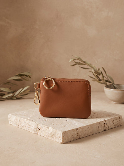 Mini Zip Coin Purse in cognac vegan leather with a key ring clip on a travertine block
