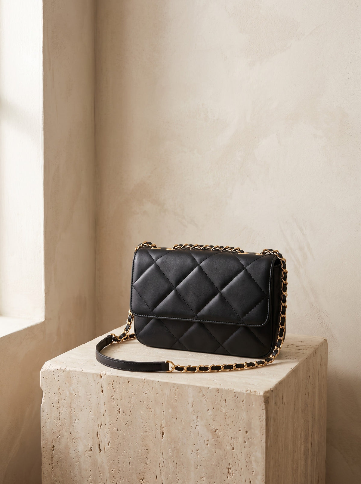 Quilted Crossbody Bag in black vegan leather with a gold-tone chain strap on a travertine plinth