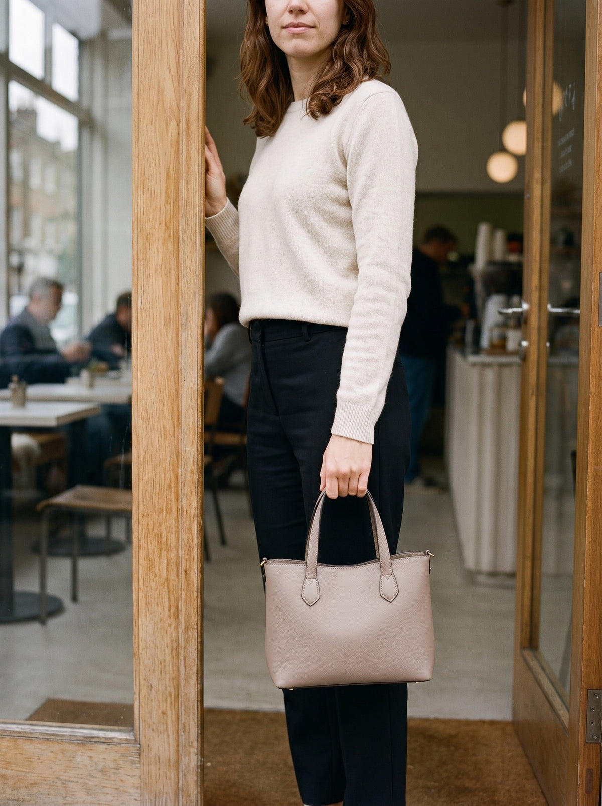 Woman holding the taupe Structured Mini Tote by its top handles in a cafe doorway